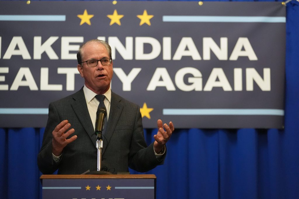 Indiana Governor Mike Braun gestures with both hands while speaking from behind a podium at a "Make Indiana Healthy Again" initiative event. He is wearing a dark grey suit jacket over a white dress shirt, a dark blue patterned tie and glasses.