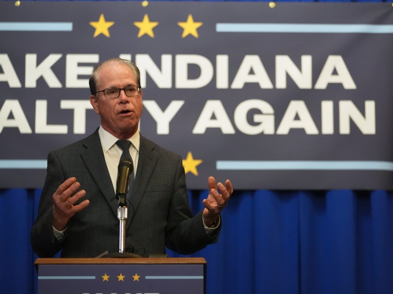 Indiana Governor Mike Braun gestures with both hands while speaking from behind a podium at a "Make Indiana Healthy Again" initiative event. He is wearing a dark grey suit jacket over a white dress shirt, a dark blue patterned tie and glasses.