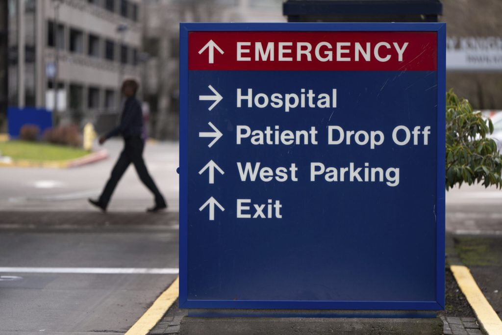 A hospital directional sign reads "EMERGENCY," "Hospital," "Patient Drop Off," "West Parking," and "Exit."