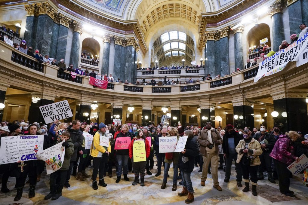 A large crowd of demonstrators gathers inside a grand, multi-tiered capitol rotunda holding pro-choice protest signs, including one that reads "Roe, Roe, Roe Your Vote." The attendees are dressed in a variety of casual winter clothing, including heavy coats, parkas, beanies and scarves.