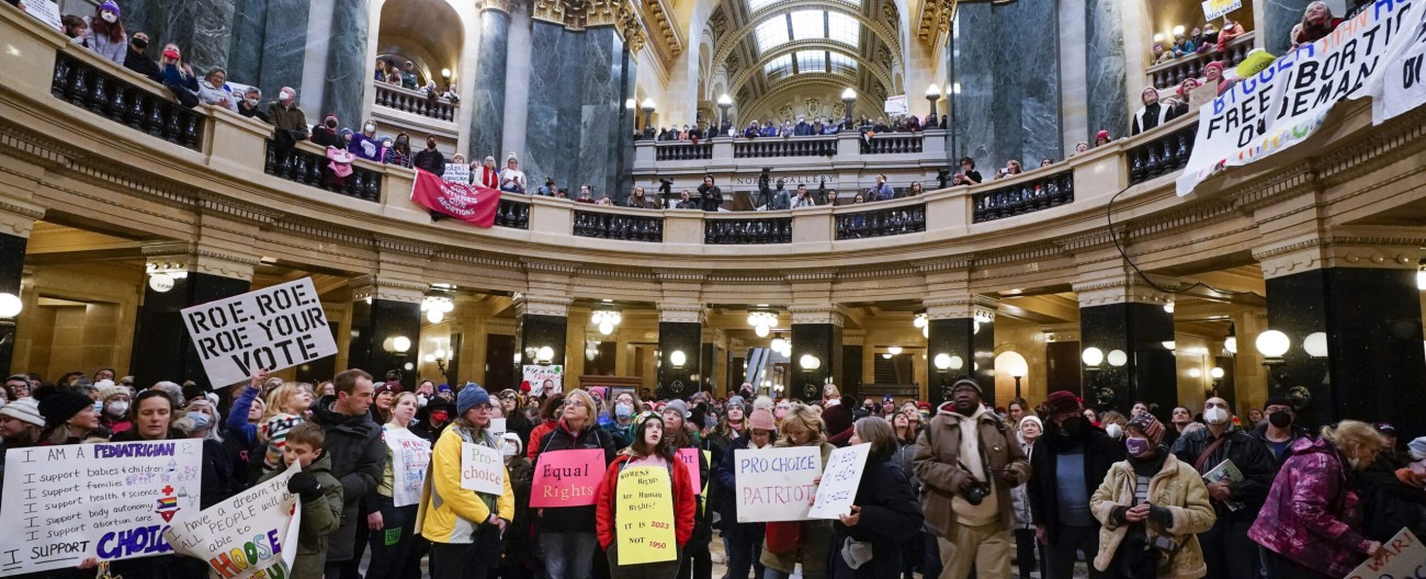 A large crowd of demonstrators gathers inside a grand, multi-tiered capitol rotunda holding pro-choice protest signs, including one that reads "Roe, Roe, Roe Your Vote." The attendees are dressed in a variety of casual winter clothing, including heavy coats, parkas, beanies and scarves.
