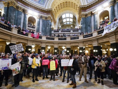 A large crowd of demonstrators gathers inside a grand, multi-tiered capitol rotunda holding pro-choice protest signs, including one that reads "Roe, Roe, Roe Your Vote." The attendees are dressed in a variety of casual winter clothing, including heavy coats, parkas, beanies and scarves.