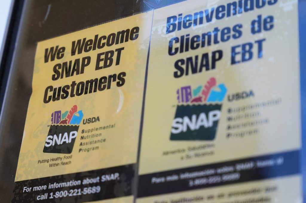 Two signs are posted side-by-side on a glass surface welcoming SNAP EBT customers, with one written in English and the other in Spanish. Both signs feature the official USDA Supplemental Nutrition Assistance Program logo alongside a toll-free informational phone number printed at the bottom.