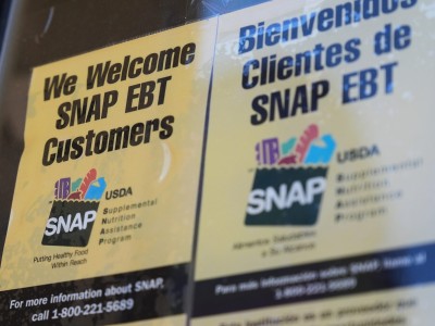 Two signs are posted side-by-side on a glass surface welcoming SNAP EBT customers, with one written in English and the other in Spanish. Both signs feature the official USDA Supplemental Nutrition Assistance Program logo alongside a toll-free informational phone number printed at the bottom.
