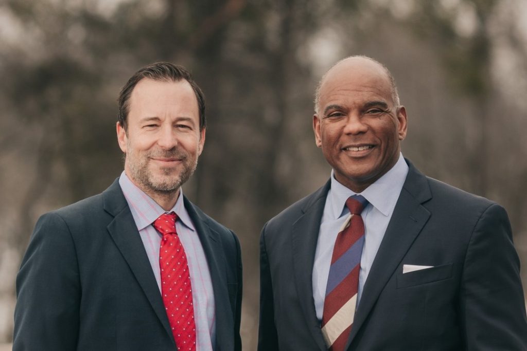 A medium close-up shot shows two men, Brian Nicholson on the left and Kendall Qualls on the right, standing side by side and smiling against a soft, blurred outdoor background. The man on the left wears a dark suit jacket, a light patterned button-down shirt, and a red polka-dot tie, while the man on the right wears a dark suit with a light blue shirt, a striped tie and a white pocket square.