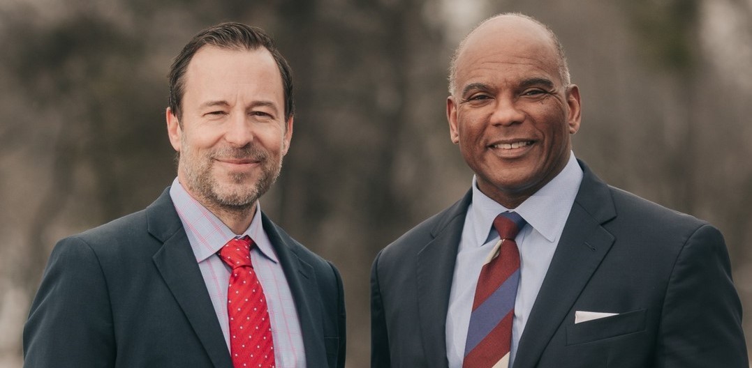 A medium close-up shot shows two men, Brian Nicholson on the left and Kendall Qualls on the right, standing side by side and smiling against a soft, blurred outdoor background. The man on the left wears a dark suit jacket, a light patterned button-down shirt, and a red polka-dot tie, while the man on the right wears a dark suit with a light blue shirt, a striped tie and a white pocket square.