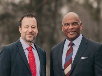 A medium close-up shot shows two men, Brian Nicholson on the left and Kendall Qualls on the right, standing side by side and smiling against a soft, blurred outdoor background. The man on the left wears a dark suit jacket, a light patterned button-down shirt, and a red polka-dot tie, while the man on the right wears a dark suit with a light blue shirt, a striped tie and a white pocket square.