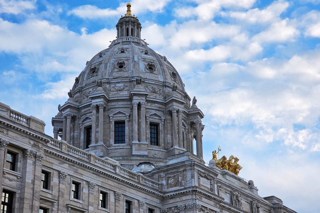 The marble dome and upper exterior of the Minnesota State Capitol building, featuring columns, arched windows and the golden Quadriga statue group.