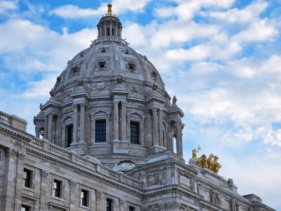The marble dome and upper exterior of the Minnesota State Capitol building, featuring columns, arched windows and the golden Quadriga statue group.