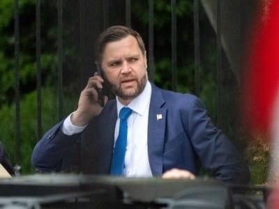 Vice President JD Vance, in a navy suit, white shirt, and bright blue tie, holds a phone to his ear while standing outdoors. He appears to be looking to the side, with a dark fence and green trees behind him. A blurred red-and-white shape partly covers the right side of the image.