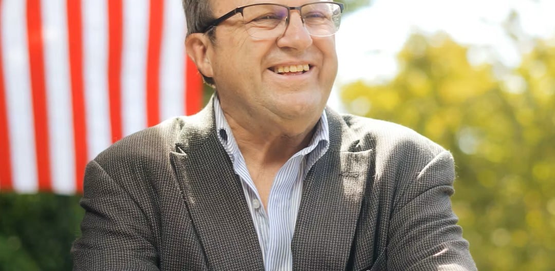 A softly-lit photo of Dean Kaufert smiling, positioned in front of an American flag and a soft-focus background of green trees. He is looking off-camera to the right with a cheerful expression.