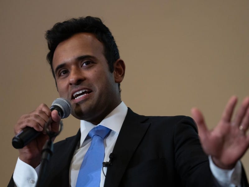A close-up shot shows Vivek Ramaswamy speaking into a microphone with his left hand raised in a gesturing motion. He is wearing a black suit jacket over a white dress shirt and a bright, light-blue necktie.