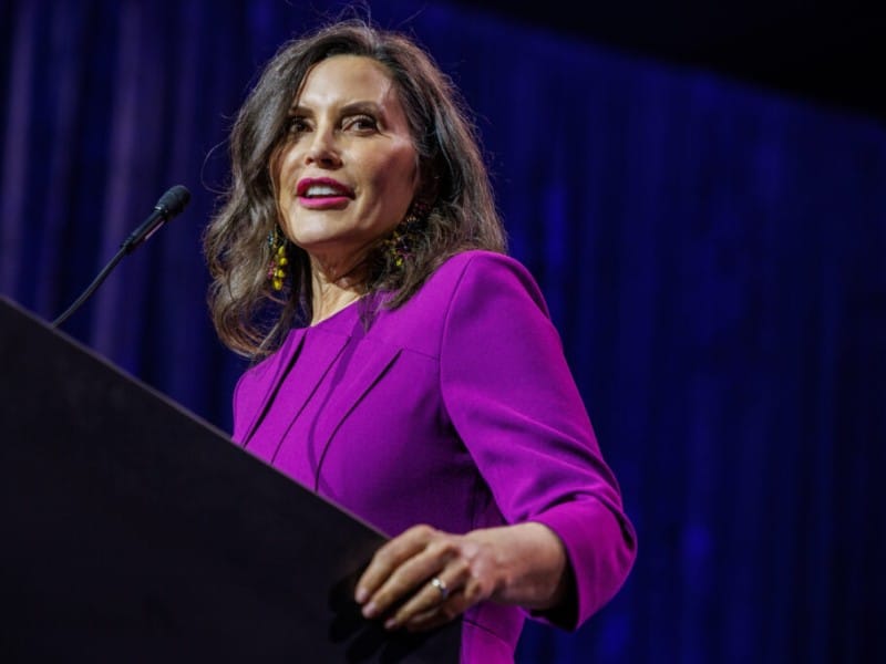 Michigan Gov. Gretchen Whitmer stands at a wooden lectern and speaks into a microphone against a dark blue curtain backdrop. She is wearing a structured magenta dress with three-quarter length sleeves, vibrant pink lipstick and large, colorful beaded earrings.
