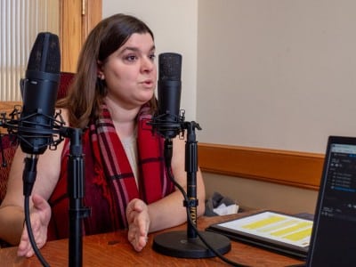 A photo of Haley Kottler from the nonpartisan advocacy organization Kansas Appleseed, a woman wearing a prominent red and black plaid scarf sits at a wooden desk, gesturing with her hands as she speaks into a black studio microphone.