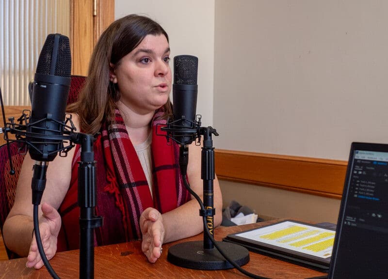 A photo of Haley Kottler from the nonpartisan advocacy organization Kansas Appleseed, a woman wearing a prominent red and black plaid scarf sits at a wooden desk, gesturing with her hands as she speaks into a black studio microphone.