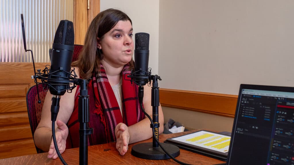 A photo of Haley Kottler from the nonpartisan advocacy organization Kansas Appleseed, a woman wearing a prominent red and black plaid scarf sits at a wooden desk, gesturing with her hands as she speaks into a black studio microphone.