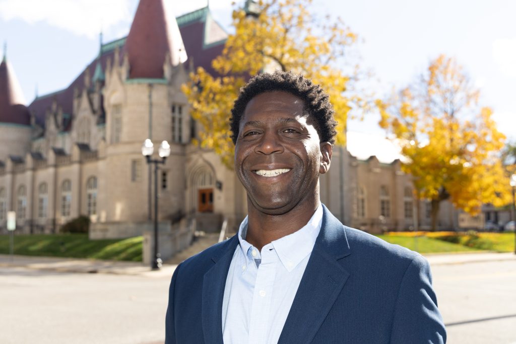 Chedrick Greene, winner of the Michigan 35th Senate District special election, smiles in front of a historic stone building with yellow autumn trees in the background
