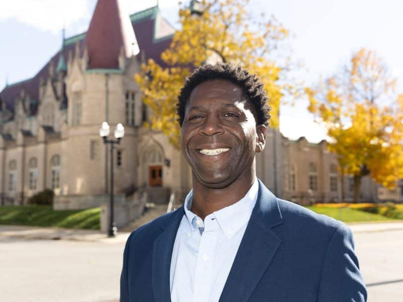 Chedrick Greene, winner of the Michigan 35th Senate District special election, smiles in front of a historic stone building with yellow autumn trees in the background
