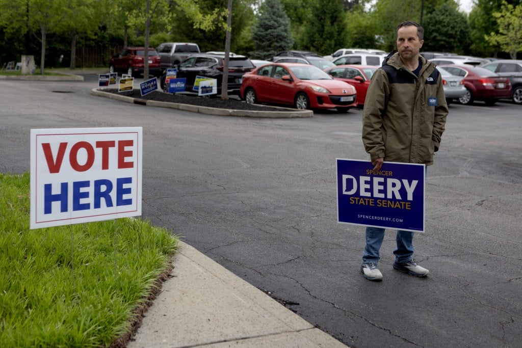 A photograph taken outdoors near a parking lot. In the left foreground, a white yard sign planted in green grass reads "VOTE HERE" in red and blue text. To the right, Indiana state Senator Spencer Deery stands holding a large campaign sign with the text "SPENCER DEERY STATE SENATE" and a website "SPENCERDEERY.COM" at the bottom. He also wears a small "DEERY" pin on his jacket. The background shows parked cars and trees.