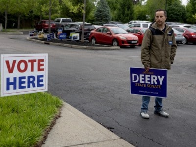 A photograph taken outdoors near a parking lot. In the left foreground, a white yard sign planted in green grass reads "VOTE HERE" in red and blue text. To the right, Indiana state Senator Spencer Deery stands holding a large campaign sign with the text "SPENCER DEERY STATE SENATE" and a website "SPENCERDEERY.COM" at the bottom. He also wears a small "DEERY" pin on his jacket. The background shows parked cars and trees.