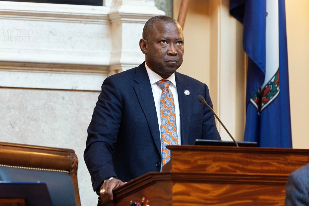 Virginia House Speaker Don Scott stands at a podium in the Virginia State Capitol. He's wearing a blue suit with a white shirt and an orange tie. He has a serious expression on his face. In the background, there is a marble wall and the state flag.
