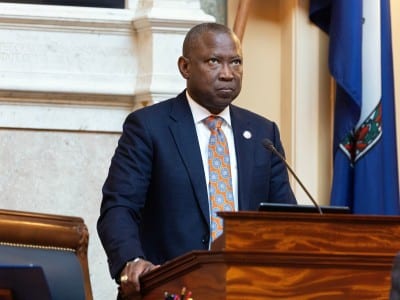 Virginia House Speaker Don Scott stands at a podium in the Virginia State Capitol. He's wearing a blue suit with a white shirt and an orange tie. He has a serious expression on his face. In the background, there is a marble wall and the state flag.