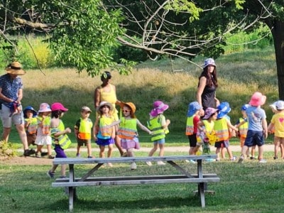 A group of diverse young children wearing neon safety vests and colorful sun hats participate in an outdoor activity in a grassy park. Several adults supervise the group near large trees and a wooden picnic table.