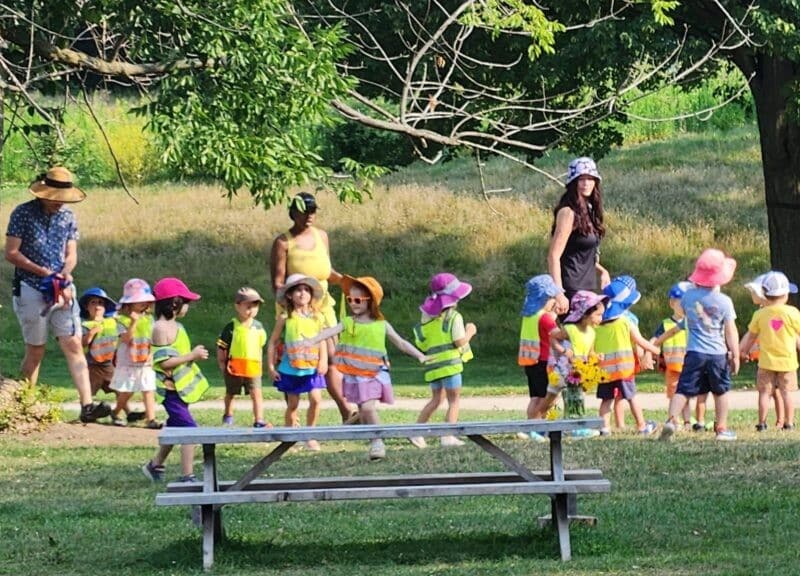 A group of diverse young children wearing neon safety vests and colorful sun hats participate in an outdoor activity in a grassy park. Several adults supervise the group near large trees and a wooden picnic table.