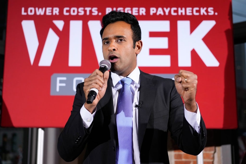 Vivek Ramaswamy speaks into a microphone with a raised fist in front of a red campaign sign that reads, “LOWER COSTS. BIGGER PAYCHECKS. VIVEK.”