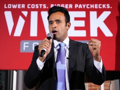 Vivek Ramaswamy speaks into a microphone with a raised fist in front of a red campaign sign that reads, “LOWER COSTS. BIGGER PAYCHECKS. VIVEK.”