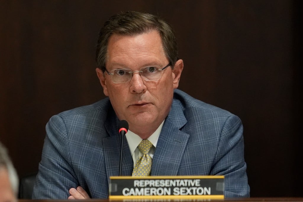 A close-up photograph of Tennessee House Speaker Cameron Sexton, a man with short brown hair and glasses, wearing a blue patterned suit and a yellow patterned tie. He is seated behind a desk with a microphone and a nameplate that reads "REPRESENTATIVE CAMERON SEXTON." The background is a solid, dark brown wall, creating a formal setting.