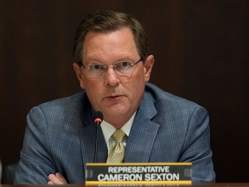 A close-up photograph of Tennessee House Speaker Cameron Sexton, a man with short brown hair and glasses, wearing a blue patterned suit and a yellow patterned tie. He is seated behind a desk with a microphone and a nameplate that reads "REPRESENTATIVE CAMERON SEXTON." The background is a solid, dark brown wall, creating a formal setting.