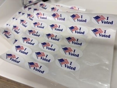 Sheets of white oval stickers feature a waving American flag graphic next to the dark blue text "I Voted."