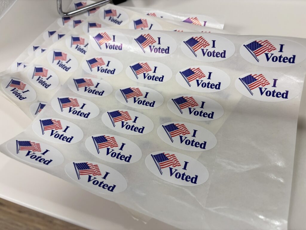 Sheets of white oval stickers feature a waving American flag graphic next to the dark blue text "I Voted."