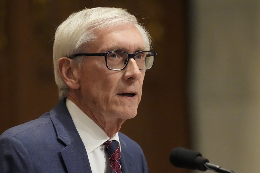 A close-up profile of Wisconsin Governor Tony Evers, an older man with short white hair and glasses, speaking into a microphone. He is wearing a blue suit jacket, a white collared shirt, and a red and navy striped tie. The background is a soft-focus, warm-toned indoor setting inside the state Capitol.
