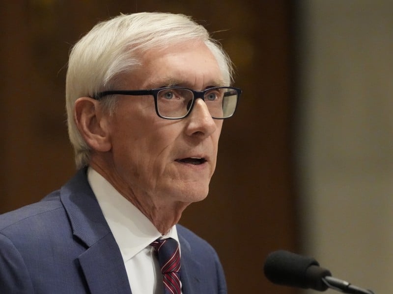 A close-up profile of Wisconsin Governor Tony Evers, an older man with short white hair and glasses, speaking into a microphone. He is wearing a blue suit jacket, a white collared shirt, and a red and navy striped tie. The background is a soft-focus, warm-toned indoor setting.