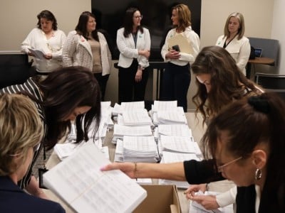 A group of women in professional attire are gathered in a bright office. They watch as staff for the North Dakota Secretary of State count petition signatures