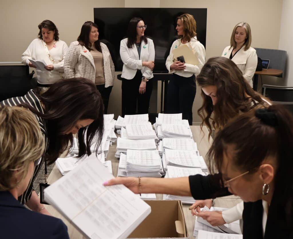 A group of women in professional attire are gathered in a bright office. They watch as staff for the North Dakota Secretary of State count petition signatures