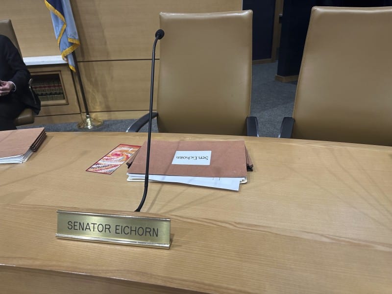 A legislative desk featuring a brass nameplate for "Senator Eichorn." The desk holds a tall microphone, a colorful pamphlet, and a stack of brown folders labeled "Sen. Eichorn" in front of two tan leather chairs.