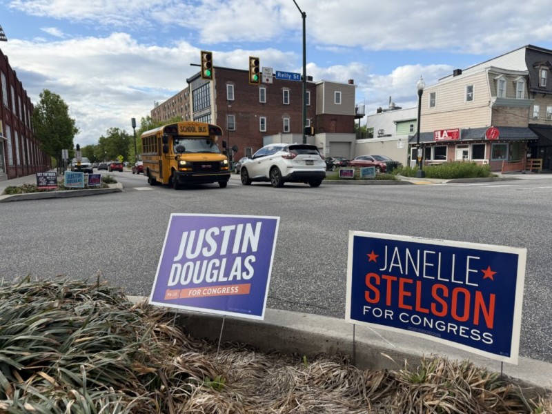 Two campaign signs for "Justin Douglas" and "Janelle Stelson" for Congress stand in the foreground of a city street corner. In the background, a yellow school bus and a white SUV drive through an intersection past brick buildings.