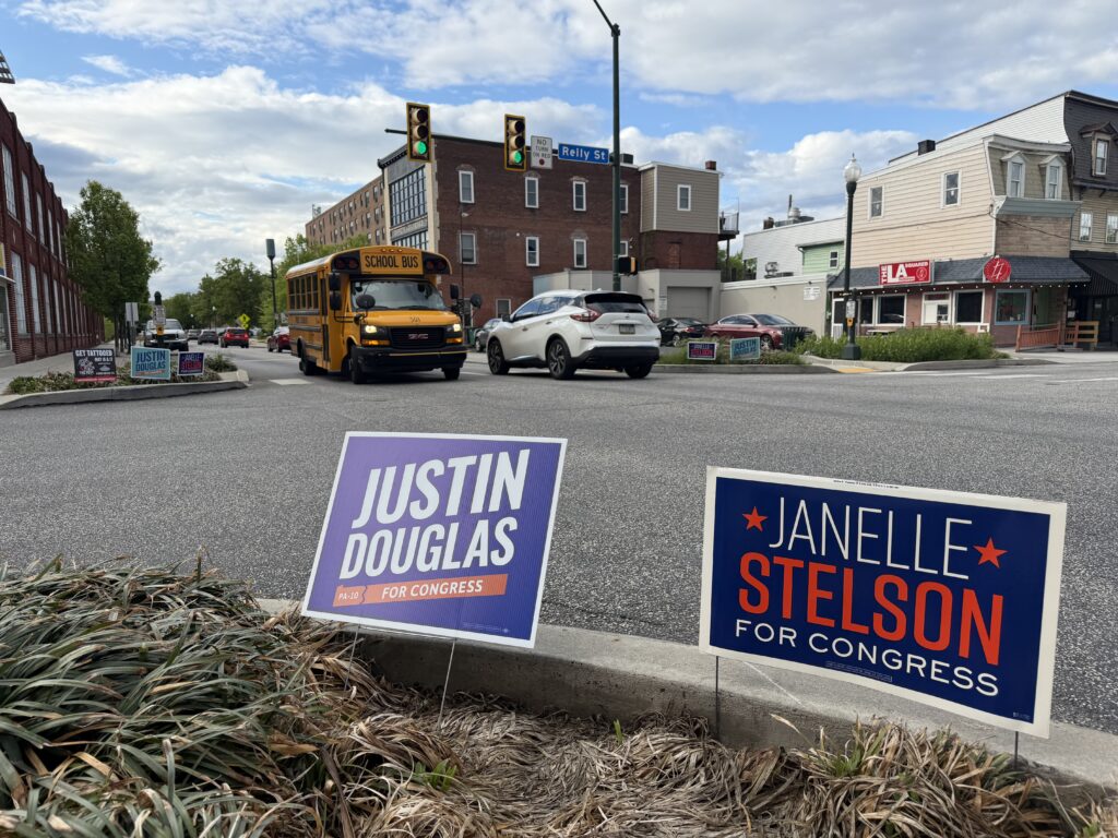 Two campaign signs for "Justin Douglas" and "Janelle Stelson" for Congress stand in the foreground of a city street corner. In the background, a yellow school bus and a white SUV drive through an intersection past brick buildings.