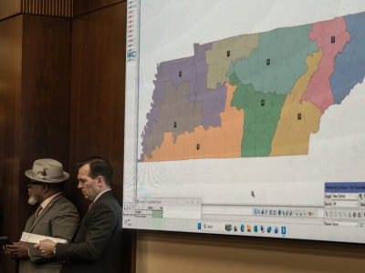 Three men in suits stand in a wood-paneled room next to a large projection screen displaying a multicolored map of Tennessee divided into legislative districts. The map features distinct regions colored in purple, orange, green, tan, pink, and blue, with single-digit numbers labeling each zone.