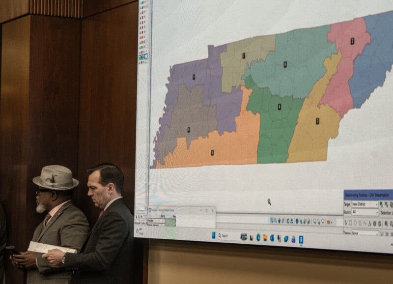 Three men in suits stand in a wood-paneled room next to a large projection screen displaying a multicolored map of Tennessee divided into legislative districts. The map features distinct regions colored in purple, orange, green, tan, pink, and blue, with single-digit numbers labeling each zone.
