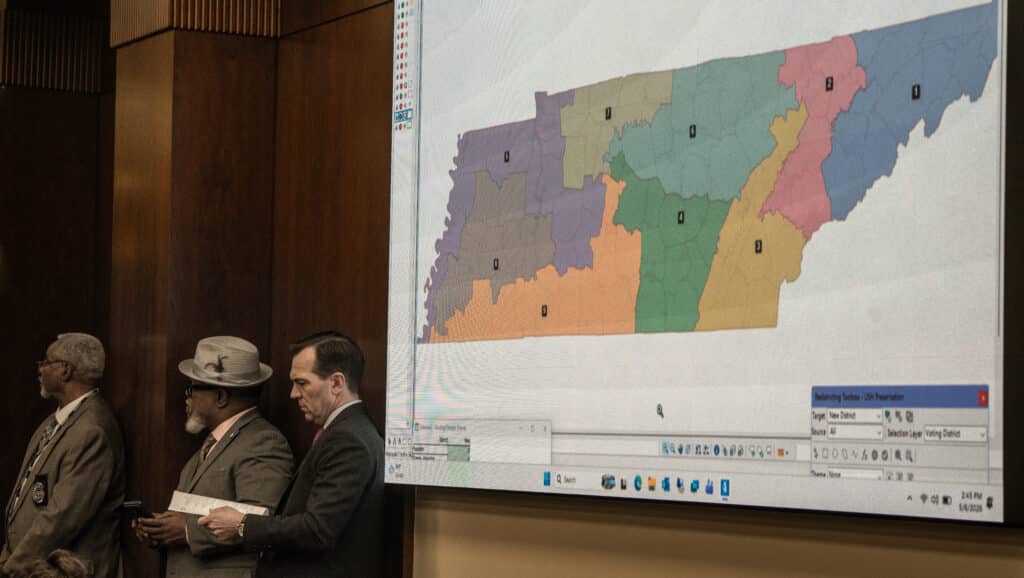 Three men in suits stand in a wood-paneled room next to a large projection screen displaying a multicolored map of Tennessee divided into legislative districts. The map features distinct regions colored in purple, orange, green, tan, pink, and blue, with single-digit numbers labeling each zone.