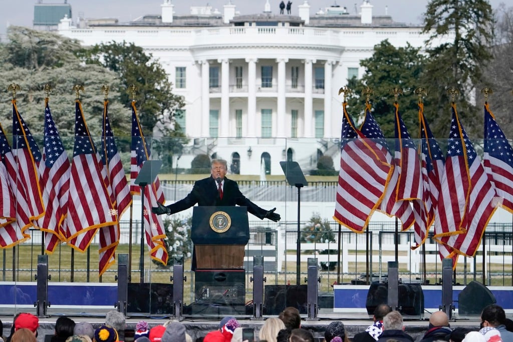 Donald Trump stands with arms outstretched at a podium bearing the presidential seal while speaking to a crowd on January 6, 2021. He is flanked by multiple red, white, and blue American flags on an outdoor stage. The White House is visible in the background.