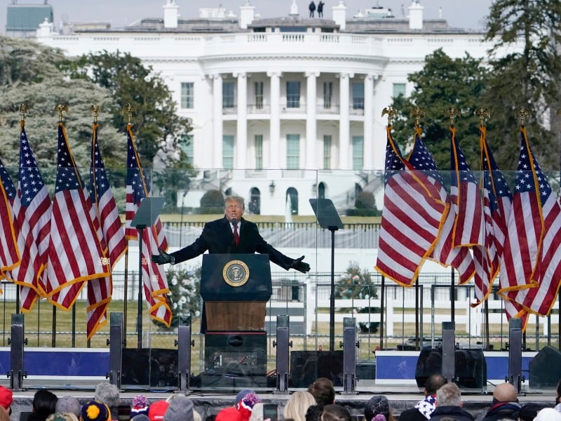 Donald Trump stands with arms outstretched at a podium bearing the presidential seal while speaking to a crowd on January 6, 2021. He is flanked by multiple red, white, and blue American flags on an outdoor stage. The White House is visible in the background.