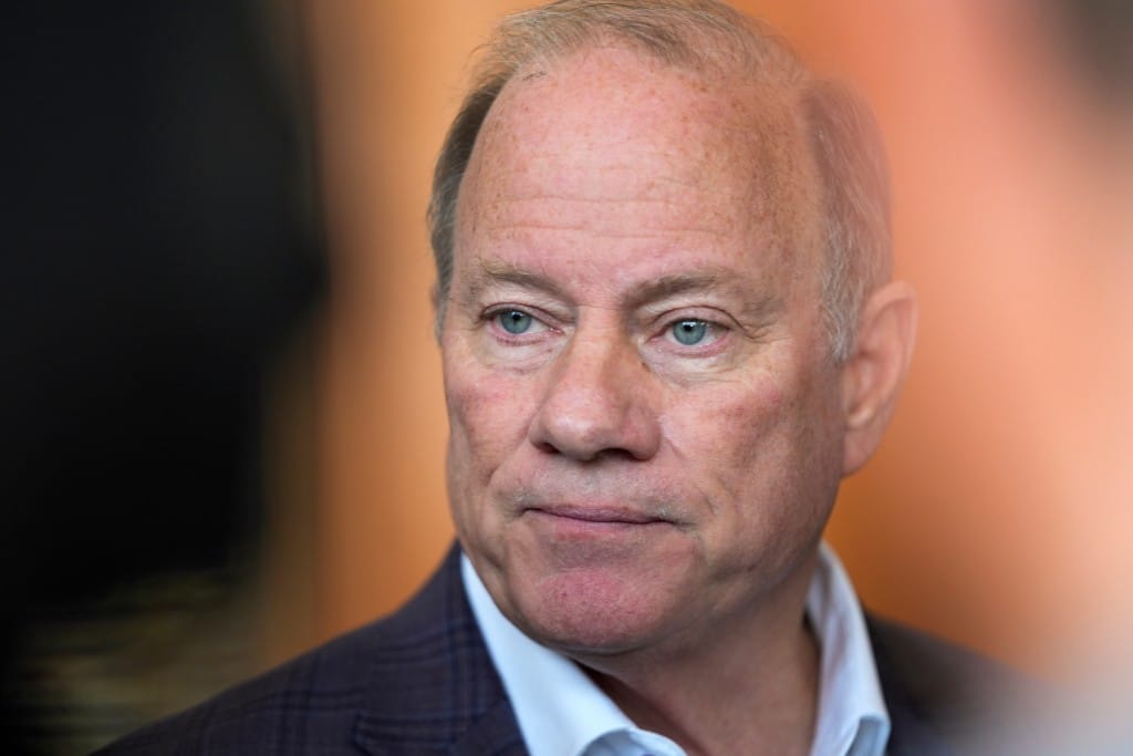A close-up portrait shows former Detroit Mayor Mike Duggan, a fair-skinned man with blue eyes and thinning light hair looking toward the side with a neutral expression. He is wearing a dark, subtly patterned blazer over an open-collared white shirt.
