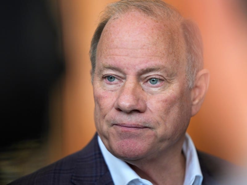 A close-up portrait shows former Detroit Mayor Mike Duggan, a fair-skinned man with blue eyes and thinning light hair looking toward the side with a neutral expression. He is wearing a dark, subtly patterned blazer over an open-collared white shirt.