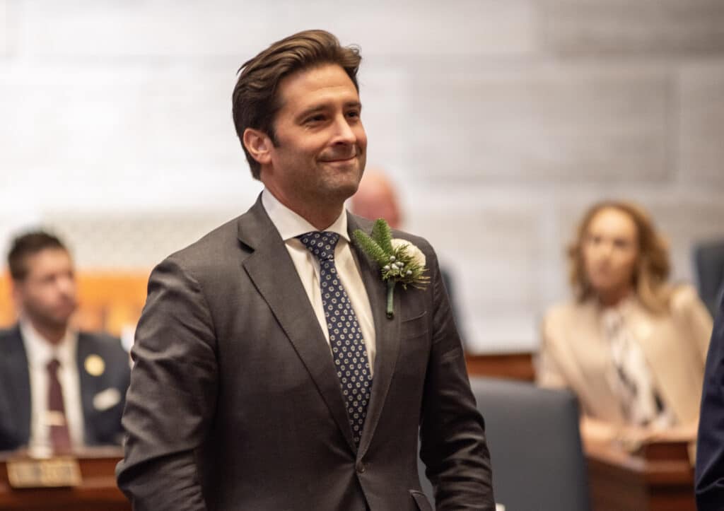 A picture of Missouri state Senator David Gregory, a man in a charcoal grey suit, white shirt, and patterned tie. He smiles while standing in a wood-paneled room. In the blurred background, other people in professional attire are seated at desks.