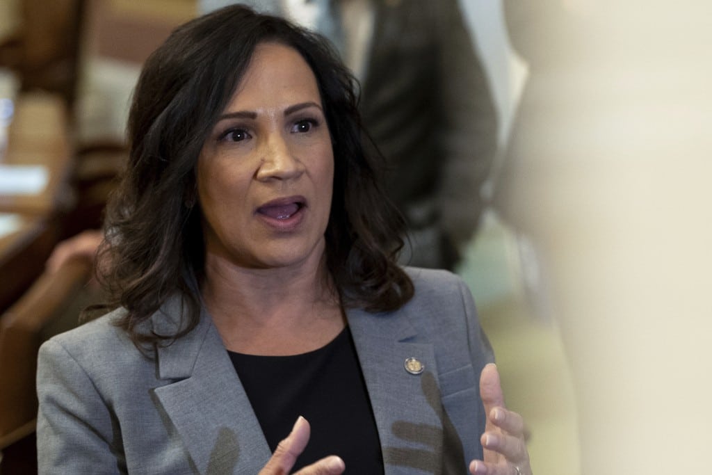An image of Minnesota House Speaker Lisa Demuth, a woman with shoulder-length, dark wavy hair speaks and gestures with both hands while wearing a gray blazer over a black top.